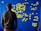 A person stands facing a blue wall covered with numerous yellow sticky notes arranged in a flowchart-like pattern. The notes are organized into categories with handwritten titles like 'Environment', 'Society', and 'Healthcare'.