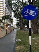 A blue bicycle sign is prominently displayed along a sidewalk lined with trees and adjacent to a street. Modern buildings with glass façades are visible on the left, alongside decorative plants. The setting conveys a peaceful urban environment suitable for cycling.