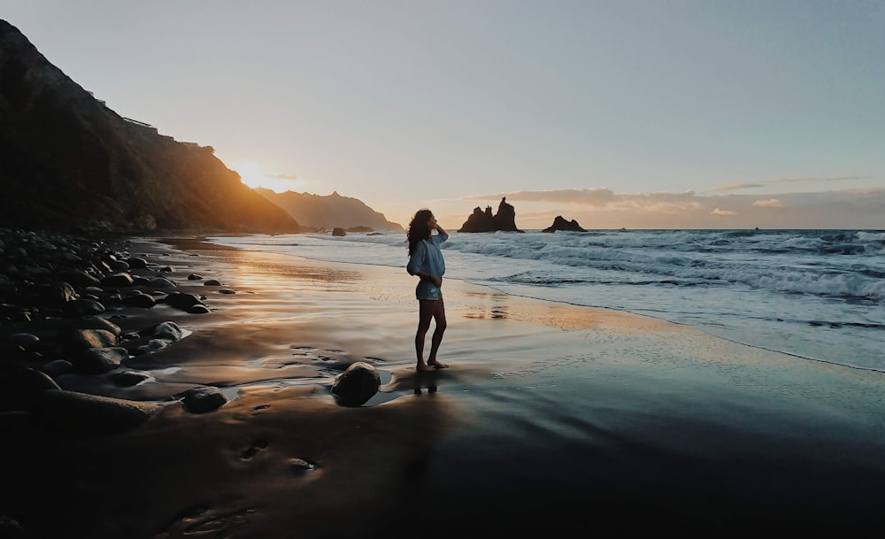 man in white shirt and black shorts standing on seashore during sunset