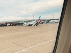 An airport tarmac with several parked airplanes. The terminal building is visible in the background, with stairs leading up to the aircraft. Passenger buses and airport service vehicles are also present.