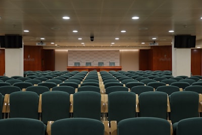 A conference room with multiple rows of green cushioned chairs facing a wooden podium with three chairs behind it. The room is well-lit with ceiling lights and features wooden paneling on the walls. Two monitors are mounted on opposite walls, and the ceiling has a grid pattern. The backdrop behind the podium displays a repeated logo.