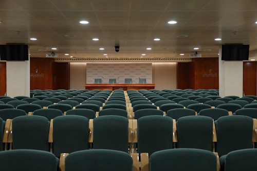 A conference room with multiple rows of green cushioned chairs facing a wooden podium with three chairs behind it. The room is well-lit with ceiling lights and features wooden paneling on the walls. Two monitors are mounted on opposite walls, and the ceiling has a grid pattern. The backdrop behind the podium displays a repeated logo.