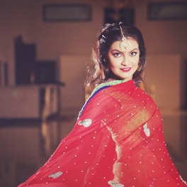 A woman with a gentle smile is adorned in a traditional red saree with intricate silver embellishments and a green blouse. She has long, dark hair and is wearing a gold maang tikka on her forehead, adding to her elegant appearance. The background is softly focused, creating a warm, intimate ambiance.