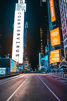 An empty urban street at night, surrounded by tall buildings with brightly lit digital billboards. The large, illuminated signs display public health messages and advertisements. The atmosphere is calm and devoid of traffic or pedestrians.