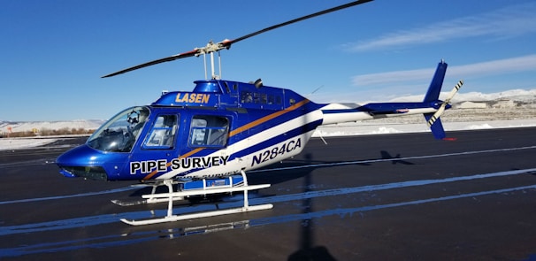 A blue and white helicopter is parked on an asphalt runway. The helicopter has 'PIPE SURVEY' written on its side, with additional text that includes 'LASEN' and 'N284CA'. The background features a clear blue sky and snow-covered mountains in the distance.