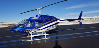 A blue and white helicopter is parked on an asphalt runway. The helicopter has 'PIPE SURVEY' written on its side, with additional text that includes 'LASEN' and 'N284CA'. The background features a clear blue sky and snow-covered mountains in the distance.