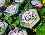 Hexagonal metal tins of face wash are placed on fresh green leaves with purple flowers scattered around. The tins have a marble design with leaf motifs and labels detailing the product as travel care items.