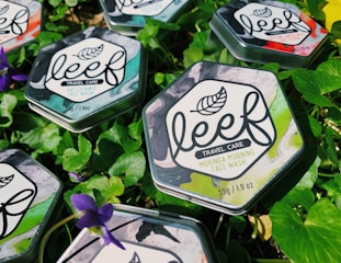 Hexagonal metal tins of face wash are placed on fresh green leaves with purple flowers scattered around. The tins have a marble design with leaf motifs and labels detailing the product as travel care items.