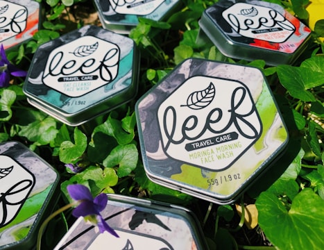 Hexagonal metal tins of face wash are placed on fresh green leaves with purple flowers scattered around. The tins have a marble design with leaf motifs and labels detailing the product as travel care items.