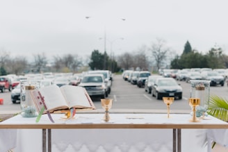 An altar setup is displayed outdoors with an open book, golden chalices, and candles on a white tablecloth against a backdrop of parked cars lining a grey parking lot. Trees and cloudy skies are visible in the distance.