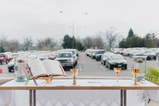 An altar setup is displayed outdoors with an open book, golden chalices, and candles on a white tablecloth against a backdrop of parked cars lining a grey parking lot. Trees and cloudy skies are visible in the distance.