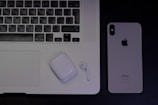 A laptop with an Arabic keyboard, a pair of wireless earphones in a charging case, and a smartphone with the Apple logo are arranged on a dark surface.