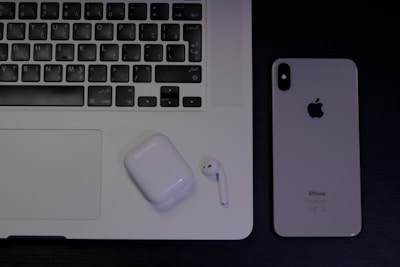 A laptop with an Arabic keyboard, a pair of wireless earphones in a charging case, and a smartphone with the Apple logo are arranged on a dark surface.