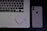 A laptop with an Arabic keyboard, a pair of wireless earphones in a charging case, and a smartphone with the Apple logo are arranged on a dark surface.