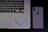 A laptop with an Arabic keyboard, a pair of wireless earphones in a charging case, and a smartphone with the Apple logo are arranged on a dark surface.