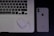 A laptop with an Arabic keyboard, a pair of wireless earphones in a charging case, and a smartphone with the Apple logo are arranged on a dark surface.