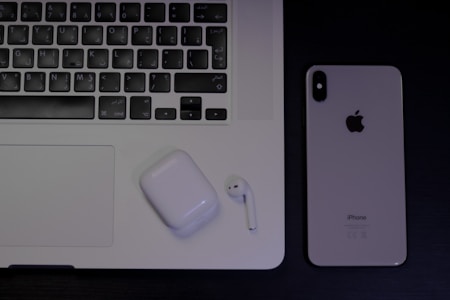 A laptop with an Arabic keyboard, a pair of wireless earphones in a charging case, and a smartphone with the Apple logo are arranged on a dark surface.