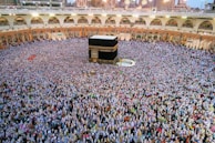 A large gathering of people surrounding the Kaaba in the Grand Mosque. The crowd appears dense, with individuals wearing a variety of light-colored clothes. The mosque's structure is visible in the background, featuring arches and multiple levels.