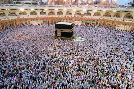 A large gathering of people surrounding the Kaaba in the Grand Mosque. The crowd appears dense, with individuals wearing a variety of light-colored clothes. The mosque's structure is visible in the background, featuring arches and multiple levels.