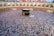 A large gathering of people surrounding the Kaaba in the Grand Mosque. The crowd appears dense, with individuals wearing a variety of light-colored clothes. The mosque's structure is visible in the background, featuring arches and multiple levels.