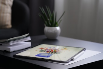 A digital tablet displaying vibrant artwork is resting on a dark-colored table. Next to the tablet is a white stylus. In the background, a small white pot holds a green plant with long, thin leaves. A stack of white notebooks or papers is partially visible on the left.