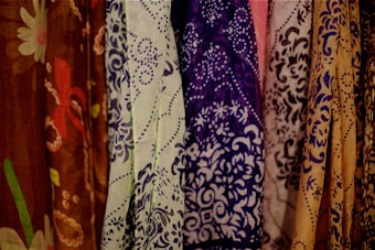 A collection of intricately patterned fabrics displayed vertically. The designs range from floral to abstract motifs on different colored fabrics, including shades of brown, green, purple, and beige.