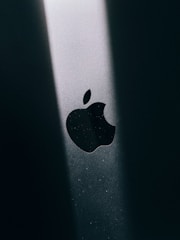 The image features a close-up of a black Apple logo, illuminated by a beam of light on a dark surface, creating a stark contrast.