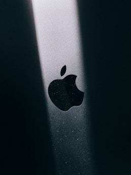 The image features a close-up of a black Apple logo, illuminated by a beam of light on a dark surface, creating a stark contrast.