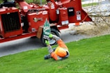A worker wearing an orange helmet and safety gear is lying on a grassy lawn holding up a sign for a tree service company. In the background, there is a large red wood chipper, and a tree branch is visible on the ground.