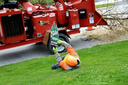 A worker wearing an orange helmet and safety gear is lying on a grassy lawn holding up a sign for a tree service company. In the background, there is a large red wood chipper, and a tree branch is visible on the ground.