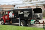 A red utility truck is equipped for tree maintenance, featuring detailed contact information and services such as lot clearing, hydro line trimming, and tree removal. The truck's design includes a hydraulic lift arm, positioned beside a residential area with green grass in the foreground.
