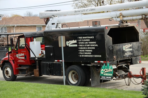 A red utility truck is equipped for tree maintenance, featuring detailed contact information and services such as lot clearing, hydro line trimming, and tree removal. The truck's design includes a hydraulic lift arm, positioned beside a residential area with green grass in the foreground.
