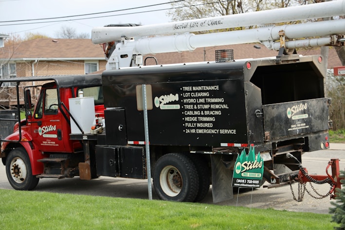 A red utility truck is equipped for tree maintenance, featuring detailed contact information and services such as lot clearing, hydro line trimming, and tree removal. The truck's design includes a hydraulic lift arm, positioned beside a residential area with green grass in the foreground.