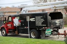 A red utility truck is equipped for tree maintenance, featuring detailed contact information and services such as lot clearing, hydro line trimming, and tree removal. The truck's design includes a hydraulic lift arm, positioned beside a residential area with green grass in the foreground.