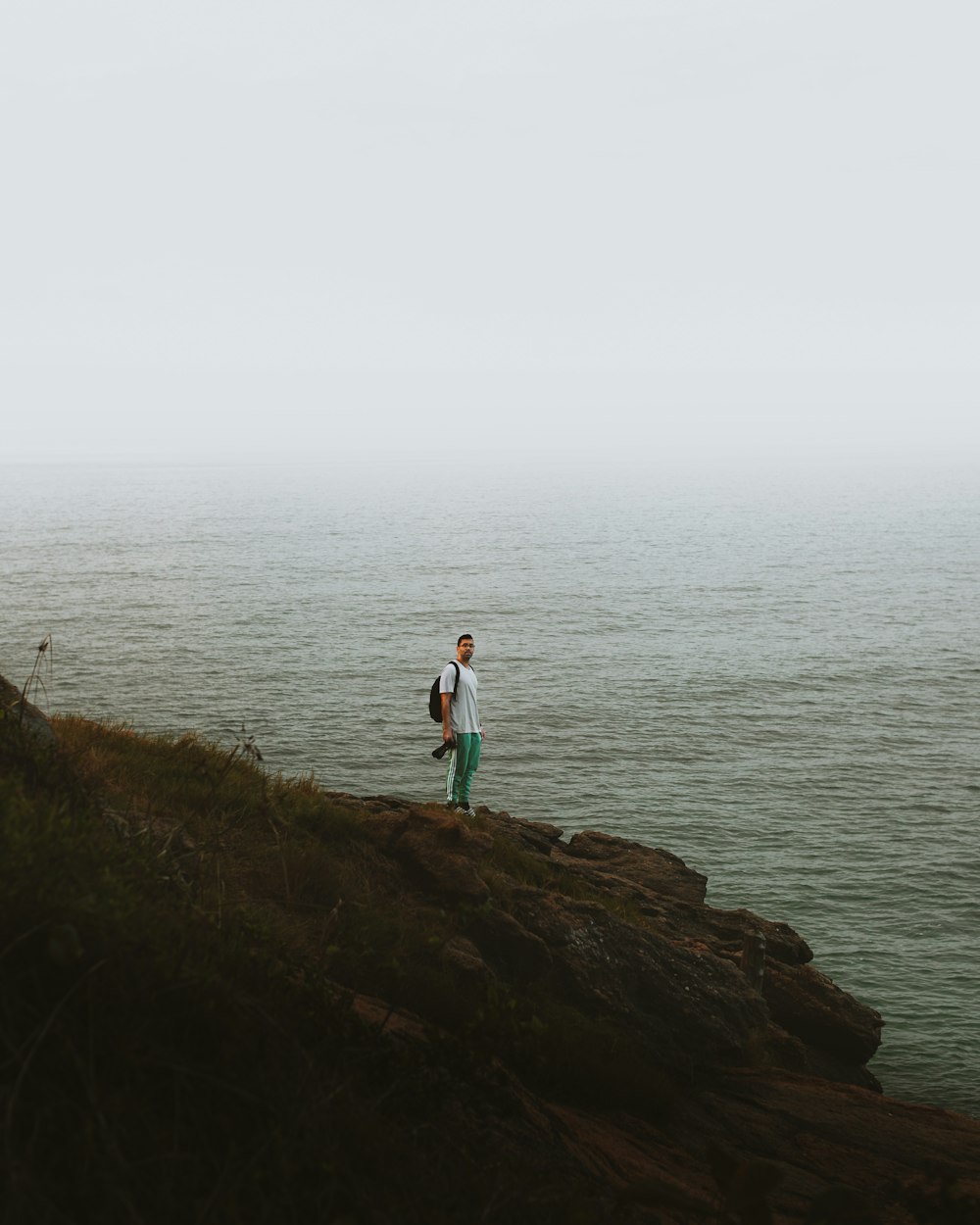 man in white t-shirt standing on brown rock near body of water during daytime