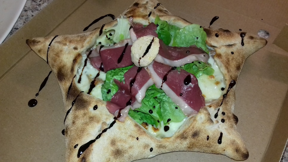 A unique, star-shaped pizza with folded corners and filled with layers of fresh lettuce, slices of cured meat, cheese, and drizzled with balsamic glaze. The crust is golden-brown and appears crisp.