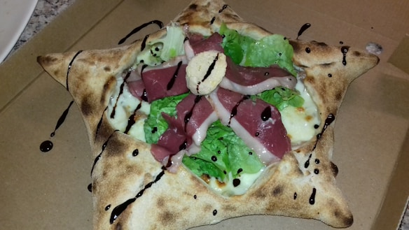 A unique, star-shaped pizza with folded corners and filled with layers of fresh lettuce, slices of cured meat, cheese, and drizzled with balsamic glaze. The crust is golden-brown and appears crisp.