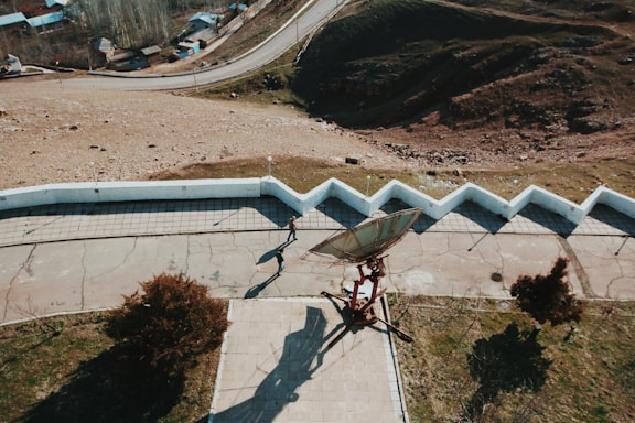 An overhead view displays a large satellite dish mounted on a platform, casting a distinct shadow on the pavement. A person walks near the dish, and a series of zigzagging white walls border a paved area. The scene is set in a hilly, semi-arid landscape with scattered trees and a few small houses in the background.