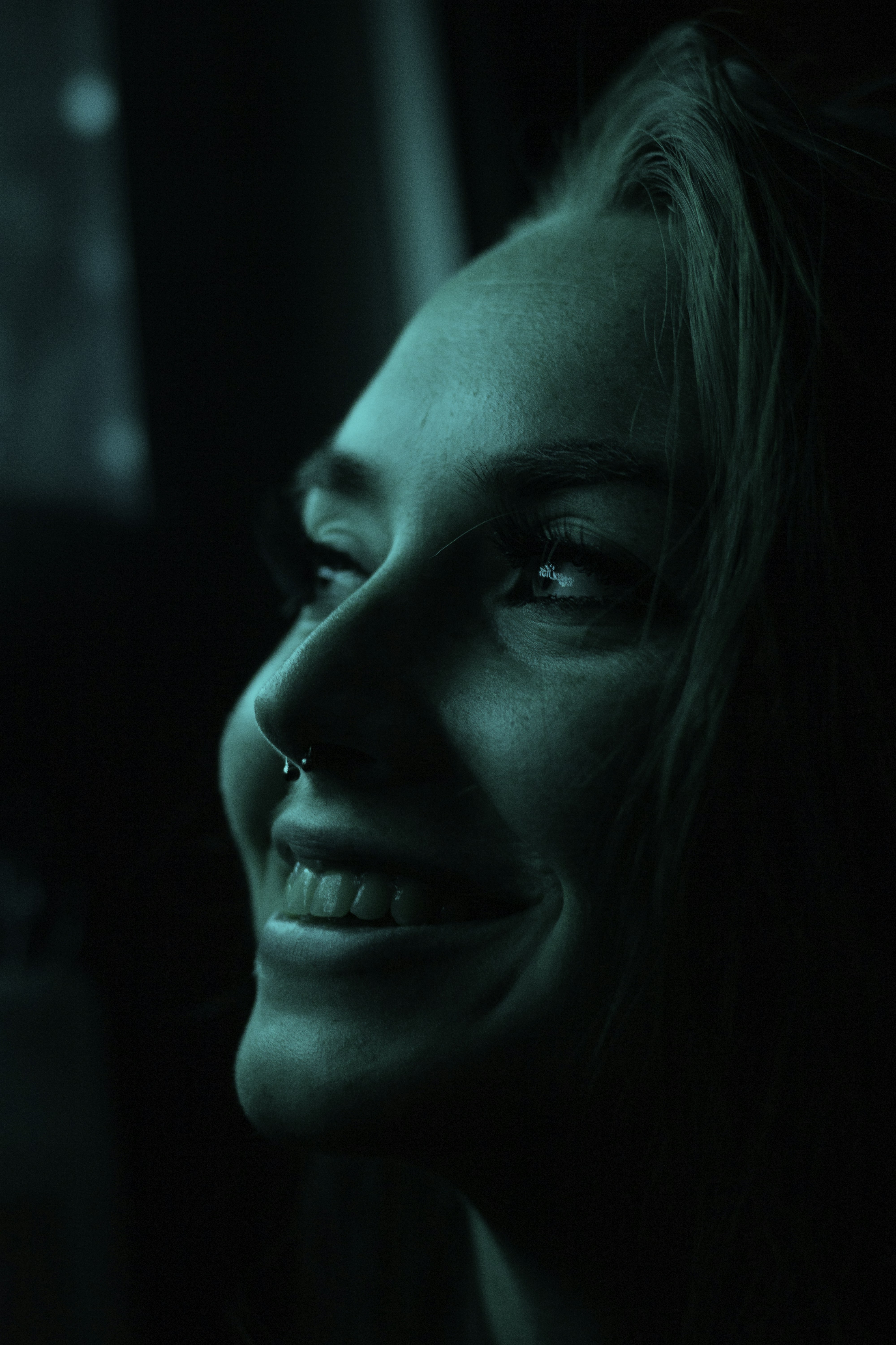 Close-up portrait photograph illuminated by cyan neon light, highlighting the subject's smile and facial features. The teal glow creates a moody, intimate atmosphere.