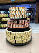 A three-tiered display stand in a store features various packaged boxes, likely food or beverage items. The top tier has boxes with white and yellow packaging, the middle tier displays brown and orange boxes, while the bottom tier shows rows of green and white boxes. In the background, there are more products on shelves and a few people are visible shopping.