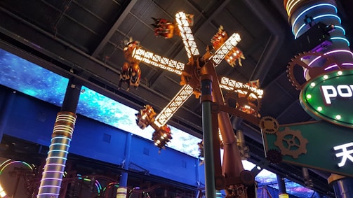 A futuristic amusement park ride is captured, with spinning mechanical arms decorated with lights. The ceiling features an illuminated space pattern, adding to the sci-fi theme. Colorful pillars and gears contribute to the mechanical, industrial vibe.