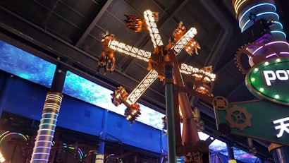 A futuristic amusement park ride is captured, with spinning mechanical arms decorated with lights. The ceiling features an illuminated space pattern, adding to the sci-fi theme. Colorful pillars and gears contribute to the mechanical, industrial vibe.