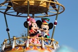 Two cartoon characters are standing on an ornate balcony decorated with colorful patterns and tassels. They are wearing festive outfits, one with a pink dress and the other with a colorful hat.