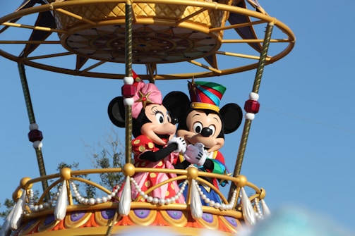 Two cartoon characters are standing on an ornate balcony decorated with colorful patterns and tassels. They are wearing festive outfits, one with a pink dress and the other with a colorful hat.