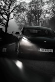 A sleek electric car with illuminated headlights is driving on a foggy, winding road. The surrounding trees are bare, adding an eerie atmosphere to the scene.