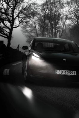 A sleek electric car with illuminated headlights is driving on a foggy, winding road. The surrounding trees are bare, adding an eerie atmosphere to the scene.