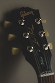 Close-up of a guitar headstock with tuning pegs and the brand name 'Gibson' prominently displayed. The background is softly blurred, enhancing the focus on the guitar's details.