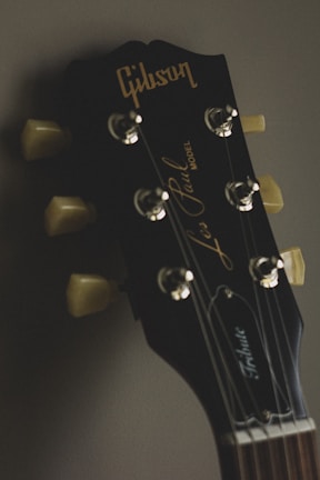 Close-up of a guitar headstock with tuning pegs and the brand name 'Gibson' prominently displayed. The background is softly blurred, enhancing the focus on the guitar's details.
