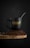 A black coffee mug with 'Fusion Coffee Beans' written on it sits atop a pile of coffee beans on a rustic wooden surface. Steam rises from the mug, indicating a hot beverage inside, and a metal spoon is placed inside the mug.