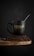 A black coffee mug with 'Fusion Coffee Beans' written on it sits atop a pile of coffee beans on a rustic wooden surface. Steam rises from the mug, indicating a hot beverage inside, and a metal spoon is placed inside the mug.